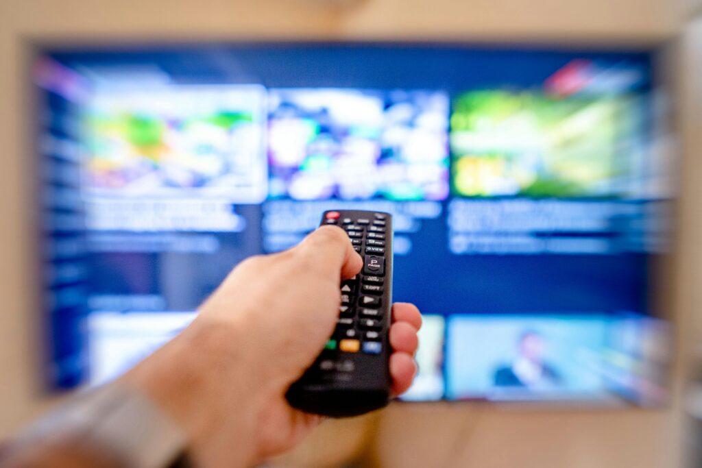 Hand holding remote control pointing at a blurred TV screen in a cozy indoor setting.